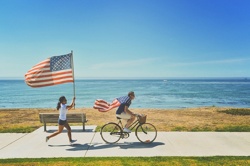 July 4th Things to do in Irvine A Man Riding a Bike with a Flag as a Cape and a Woman Running behind Him Holding a Bigger American Flag with the Ocean in the Background