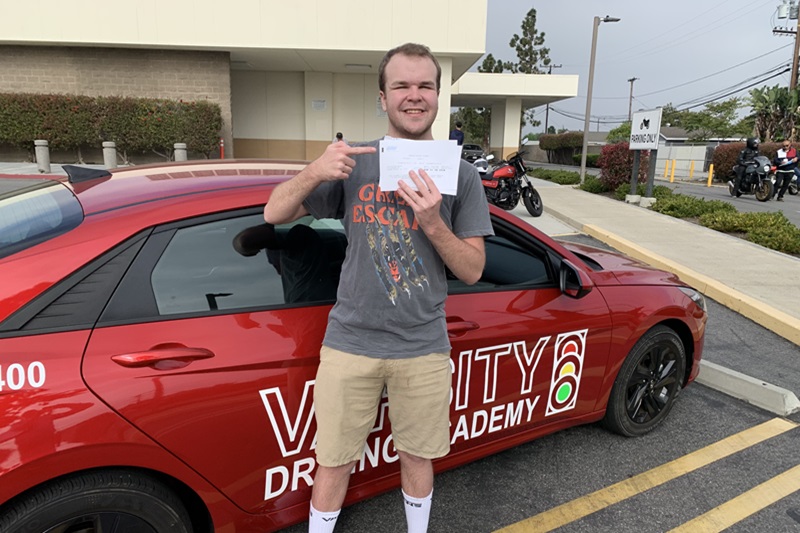 Crean Lutheran High School Driver's Ed Male Student Standing Next to a Training Vehicle Holding Up a Passed Driving Test Sheet